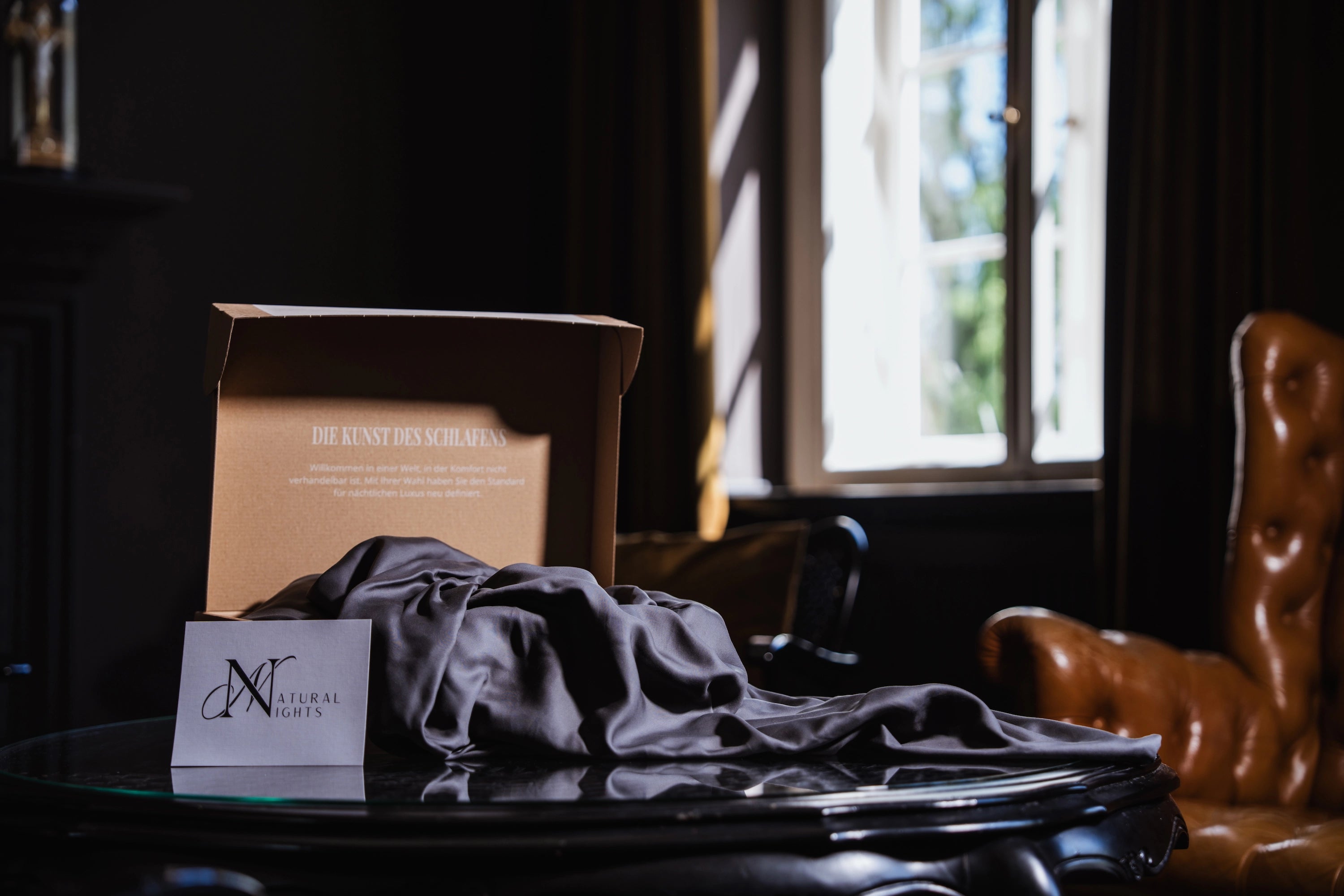 Elegante Verpackung von Natural Nights mit grauem Spannbettlaken aus TENCEL™ Lyocell auf einem runden Glastisch, im Hintergrund ein Lederstuhl und ein geöffnetes Fenster mit Tageslicht